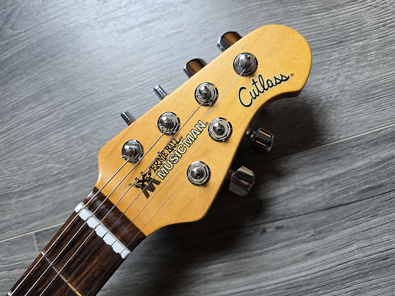 Guitar headstock with 'Ernie Ball Music Man' branding on a wooden surface