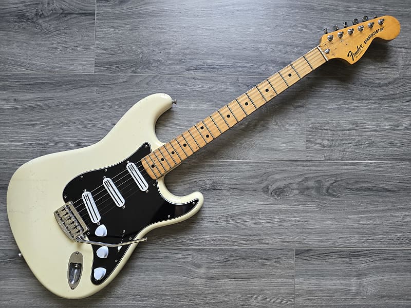 A 1979 Fender Stratocaster electric guitar in white color with a black pickguard, maple neck, and a Floyd Rose tremolo system, lying on a wooden floor.